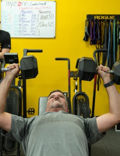 Man performing dumbbell bench press in a fitness studio, emphasizing strength training and conditioning, with a yellow wall and workout equipment in the background.