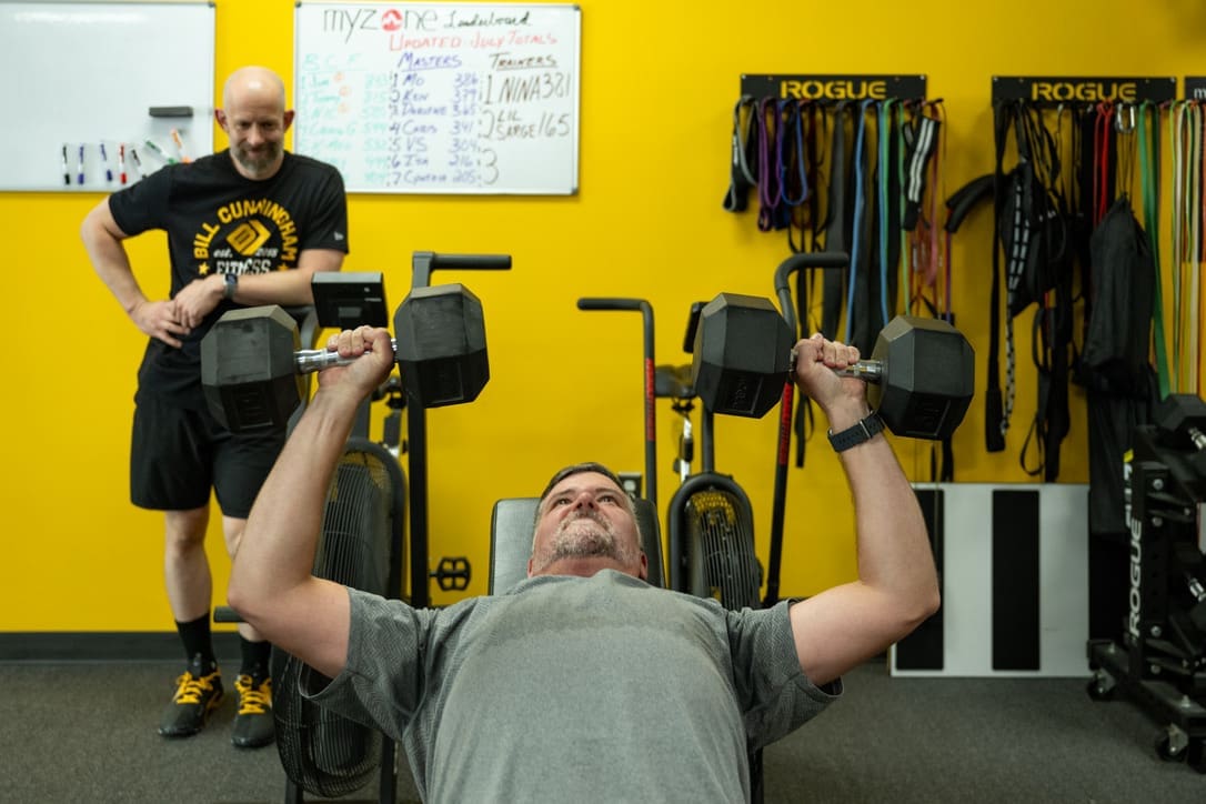 Man lifting dumbbells during small group personal training session at Bill Cunningham Fitness, with supportive trainer in background, bright yellow wall, and fitness equipment visible.