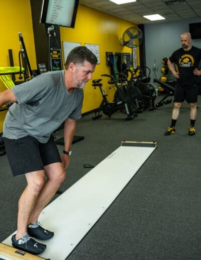 Man performing a fitness exercise on a balance board with a trainer observing in a personal training studio, emphasizing strength training and mobility improvement.