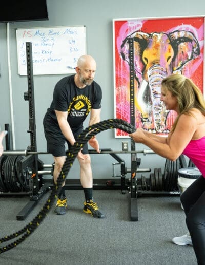 Trainer assisting a client with battle ropes workout in a gym, featuring fitness equipment and motivational artwork, showcasing personalized training at Bill Cunningham Fitness.