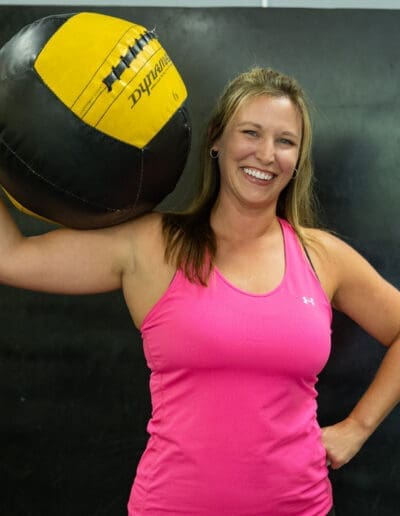 Smiling woman in a pink tank top holding a medicine ball, showcasing strength and fitness in a personal training environment.