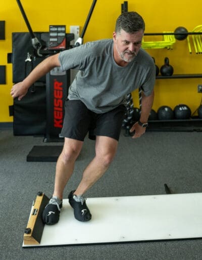 Man performing balance training on a slide board in a fitness studio, emphasizing strength and mobility improvement, with gym equipment in the background.