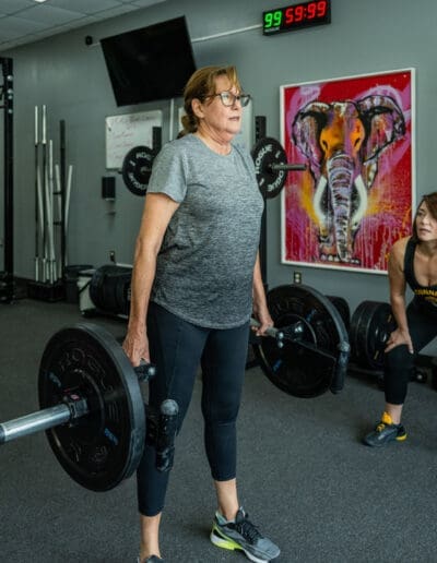 Woman performing deadlifts with a barbell in a fitness studio, showcasing strength training and personal fitness progress, with a colorful elephant mural in the background.
