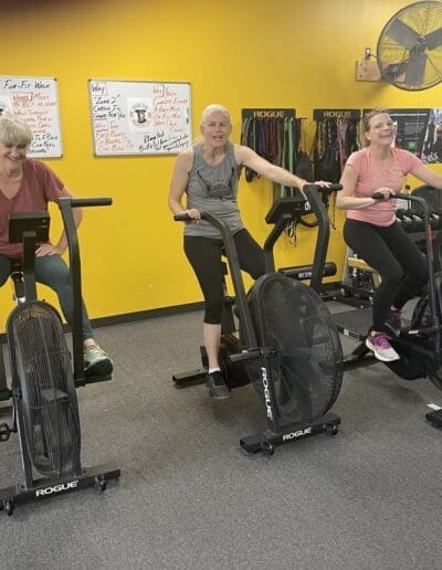 Three women engaged in a fitness session on Rogue exercise bikes, smiling and enjoying the workout in a vibrant gym environment with a yellow wall and workout equipment.