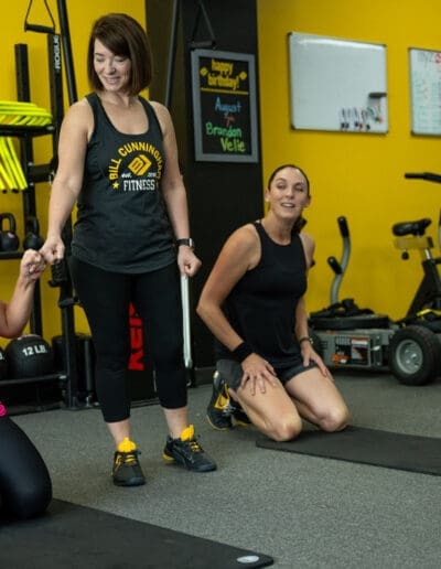 Two personal trainers from Bill Cunningham Fitness engaging with clients during a training session in a gym setting, with fitness equipment visible in the background.