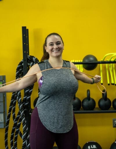 Woman smiling while demonstrating resistance band exercises in a fitness studio with yellow walls and workout equipment in the background, reflecting personal training and strength training focus.