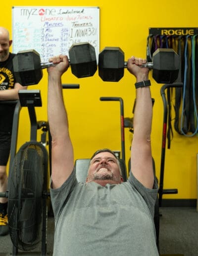 Man performing strength training exercise with dumbbells in a fitness studio, showcasing personal training at Bill Cunningham Fitness, with a yellow backdrop and fitness equipment visible.