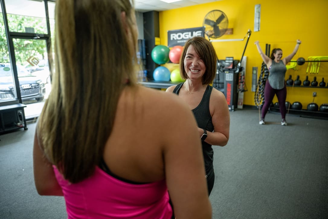 Women engaging in small group personal training, smiling and interacting in a fitness studio with exercise equipment and colorful fitness balls in the background.