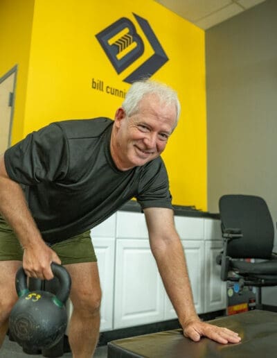 Man exercising with a kettlebell in a fitness studio, featuring the "Bill Cunningham" logo on a bright yellow wall, showcasing personal training and strength training environment.