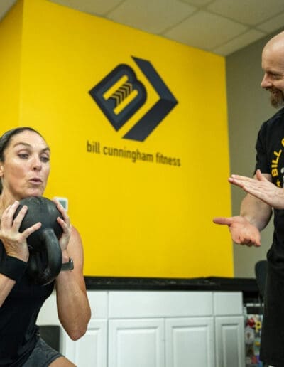Woman performing a kettlebell squat with a personal trainer providing guidance at Bill Cunningham Fitness gym.