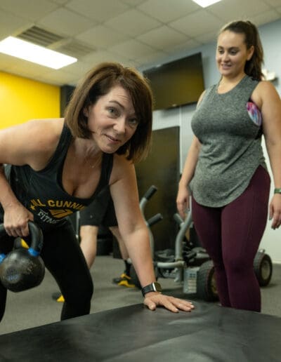Woman performing strength training with kettlebell in personal training session at Bill Cunningham Fitness, supported by another client.