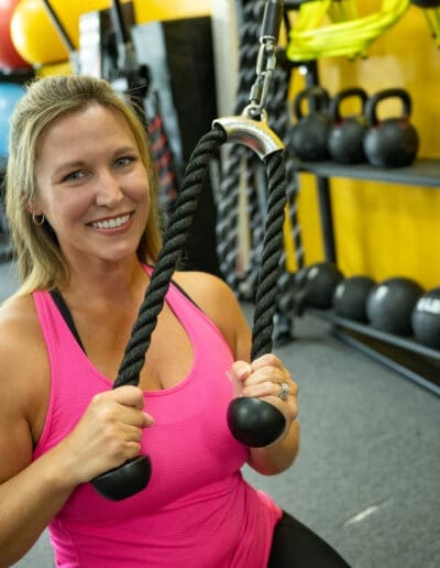 Woman in pink workout top demonstrating strength training with rope attachment in a fitness gym, featuring kettlebells in the background, emphasizing personal training and fitness transformation.