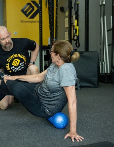 Personal trainer assisting a client with mobility exercises on a foam roller in a fitness studio, featuring Bill Cunningham Fitness branding in the background.