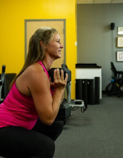 Woman performing a squat with a dumbbell in a fitness training environment, showcasing strength training and conditioning at Bill Cunningham Fitness.