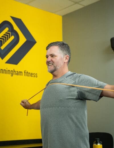 Man performing strength training exercise with resistance band in a fitness studio, featuring the Bill Cunningham Fitness logo on a vibrant yellow wall.