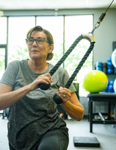 Woman exercising with cable machine in fitness studio, focusing on strength training and conditioning, surrounded by exercise equipment and fitness balls.