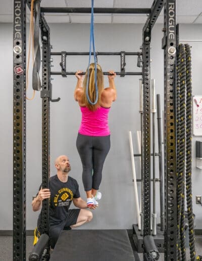 Woman performing assisted pull-ups with personal trainer in a fitness gym, showcasing strength training and personalized coaching at Bill Cunningham Fitness.