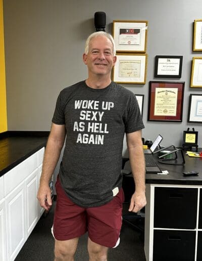 Man standing in fitness studio wearing a shirt that says "WOKE UP SEXY AS HELL AGAIN," showcasing a confident attitude in a personal training environment with fitness certifications on the wall.
