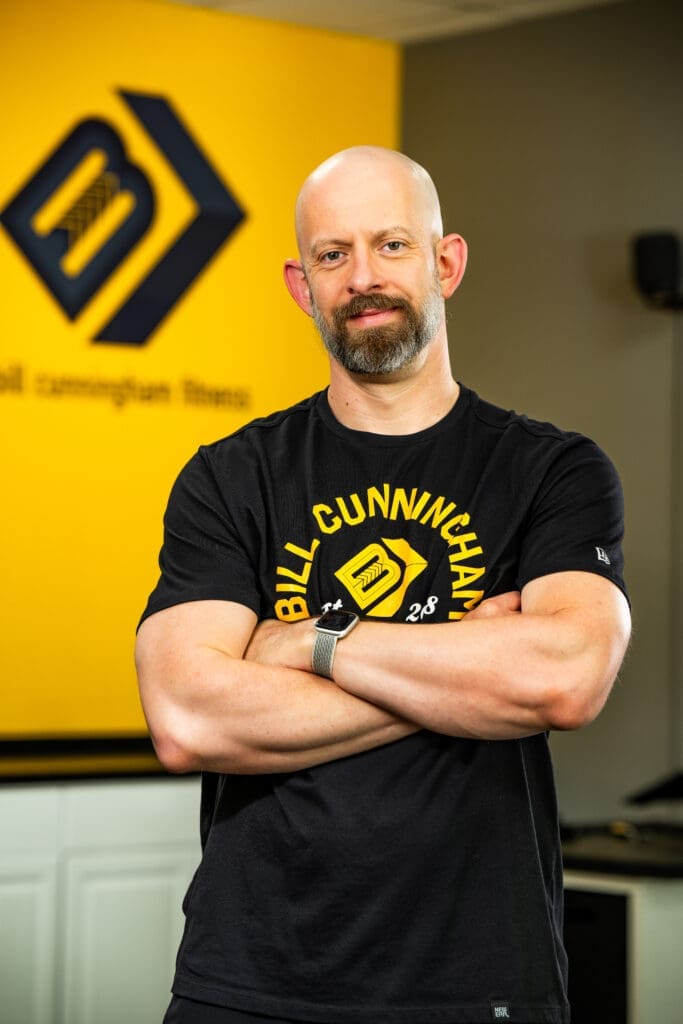 Bill Cunningham, fitness expert, standing with arms crossed, wearing a black shirt featuring "Bill Cunningham Fitness" logo, in a gym setting with a yellow wall.