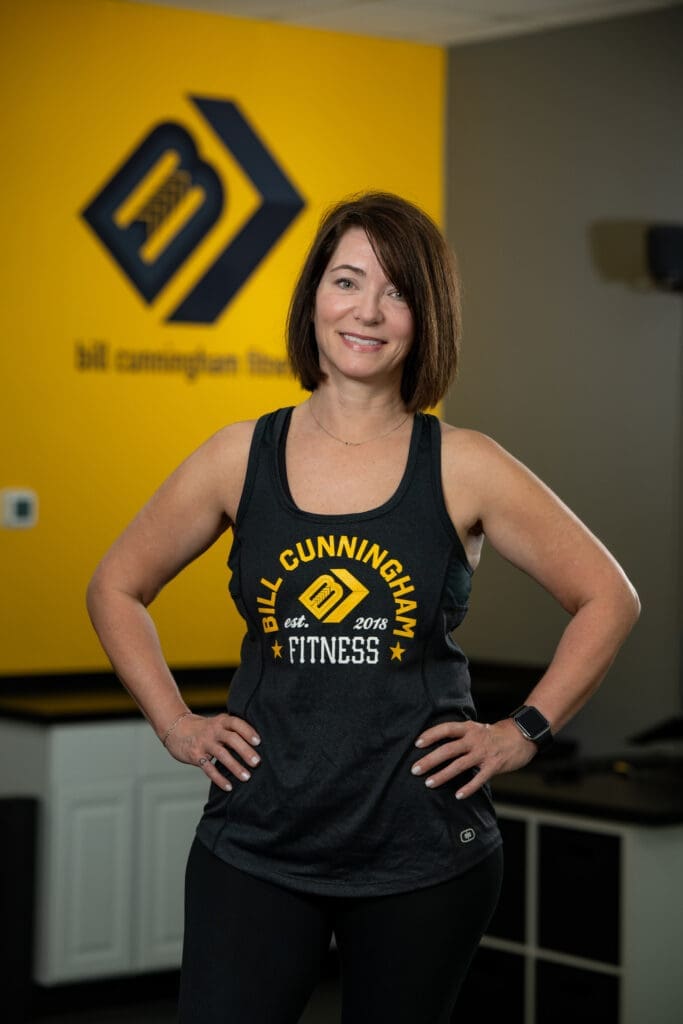 Woman in fitness attire posing confidently in front of Bill Cunningham Fitness logo, showcasing expertise in personal training and wellness coaching.